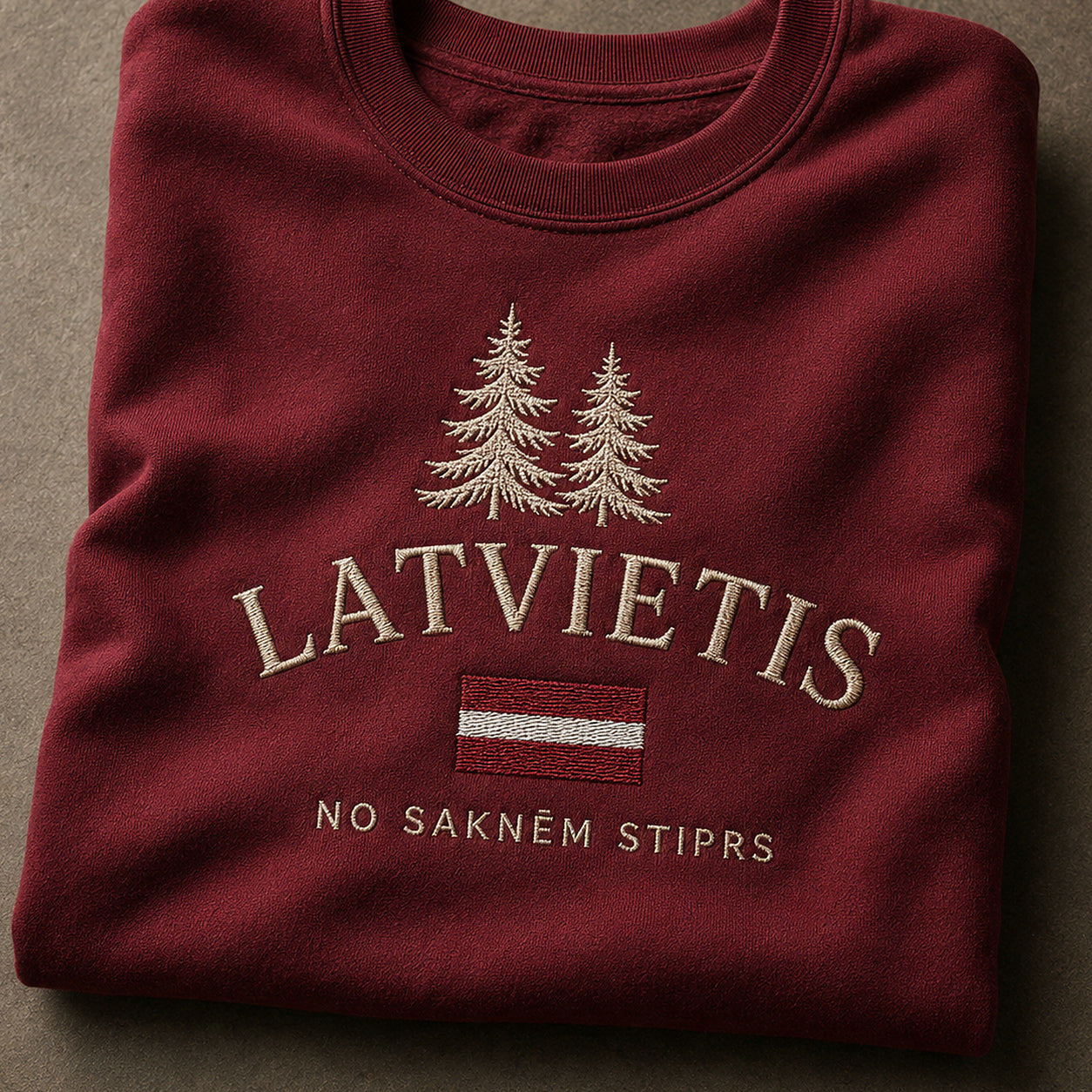 Tuvāks “Latvietis” kompozīcijas skats uz bordo salocītu džemperi ar tekstu “Latvietis” un apakštekstu “No saknēm stiprs”