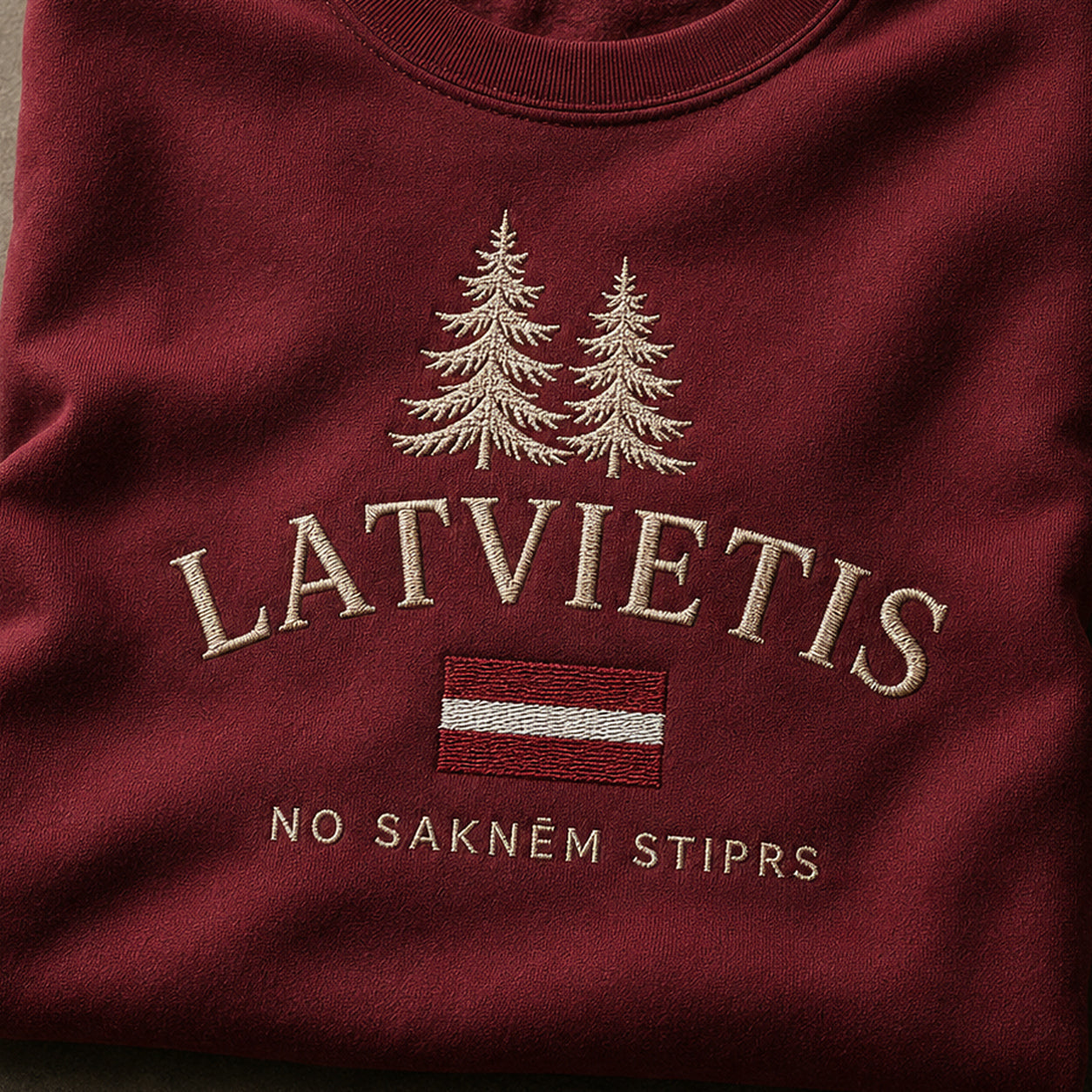 “Latvietis” izšuvuma detaļu tuvplāns uz bordo džempera auduma, redzams motīvs: lakonisks vārds Latvietis kopā ar frāzi No saknēm stiprs