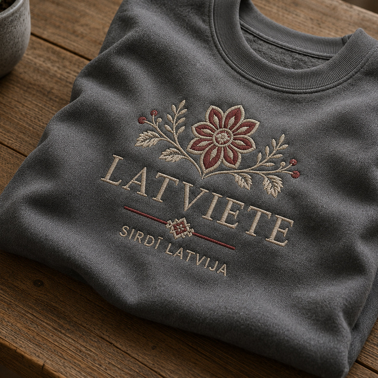 Tuvāks “Latviete” kompozīcijas skats uz pelēkas melanžas salocītu džemperi ar tekstu “Latviete” un apakštekstu “Sirdī Latvija”