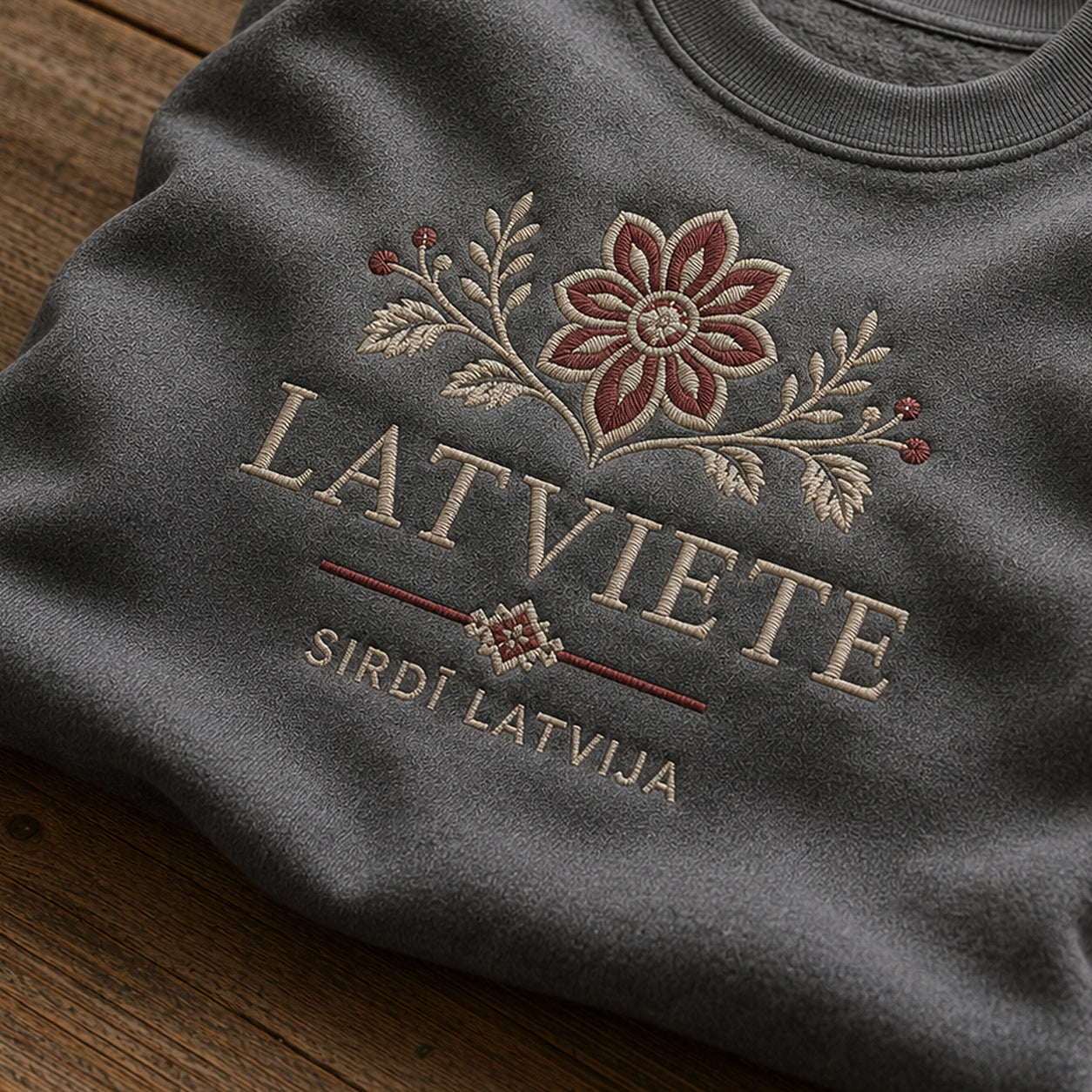 “Latviete” izšuvuma detaļu tuvplāns uz pelēkas melanžas džempera auduma, redzams motīvs: smalks uzraksts Latviete ar sirsnīgo apliecinājumu Sirdī Latvija