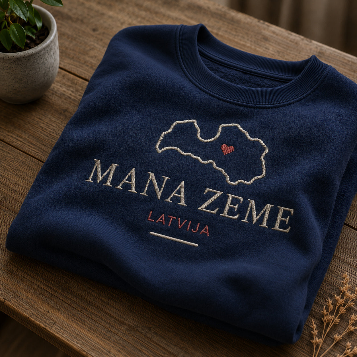 Tumši zils salocīts džemperis ar “Mana zeme Latvija” izšuvumu plašākā plakanā izkārtojuma kopskatā