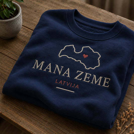 Tumši zils salocīts džemperis ar “Mana zeme Latvija” izšuvumu plašākā plakanā izkārtojuma kopskatā