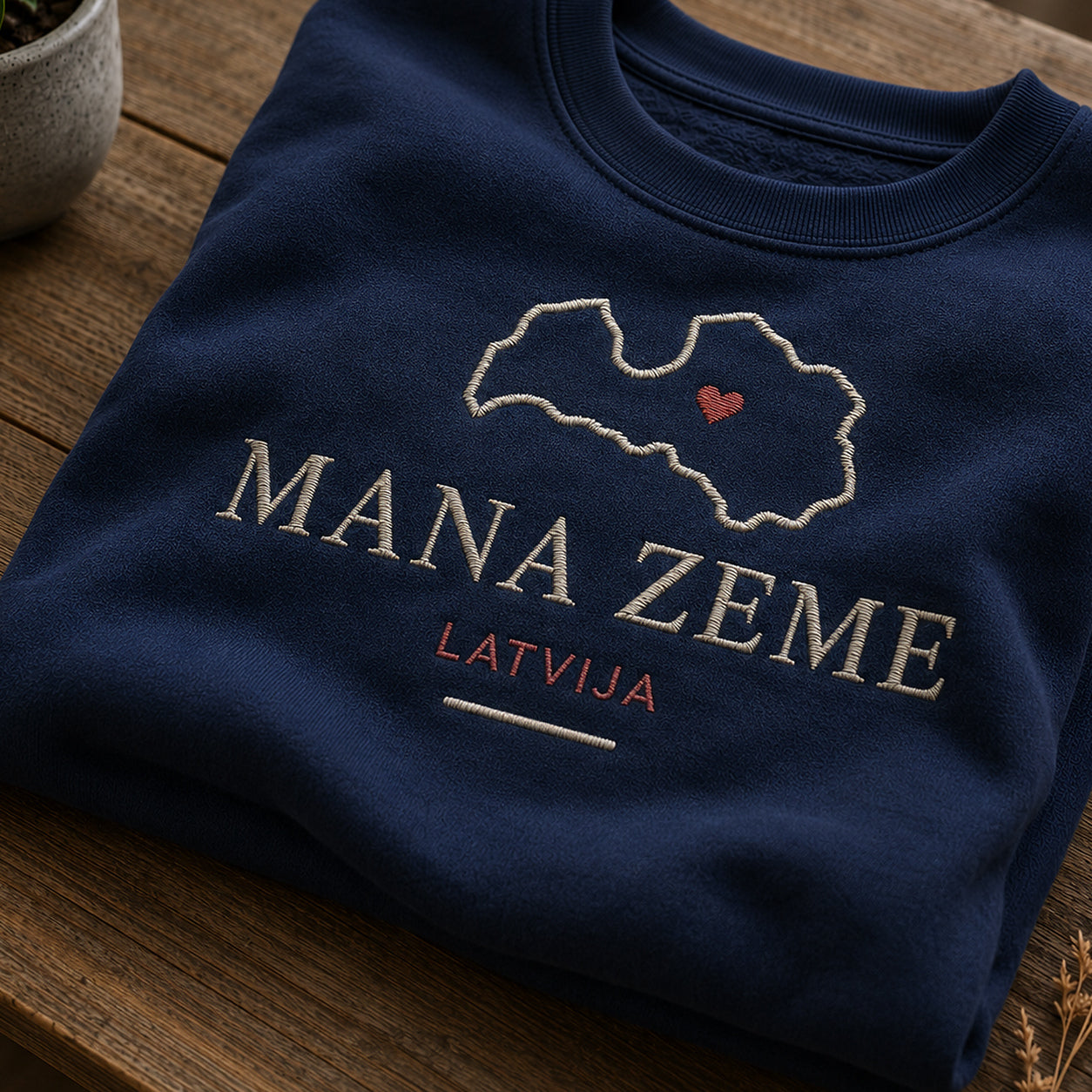 Tuvāks “Mana zeme Latvija” kompozīcijas skats uz tumši zilu salocītu džemperi ar tekstu “Mana zeme” un apakštekstu “Latvija”