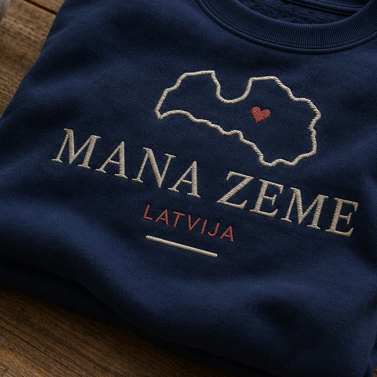 “Mana zeme Latvija” izšuvuma detaļu tuvplāns uz tumši zila džempera auduma, redzams motīvs: tiešs vēstījums Mana zeme un skaidri izcelts vārds Latvija