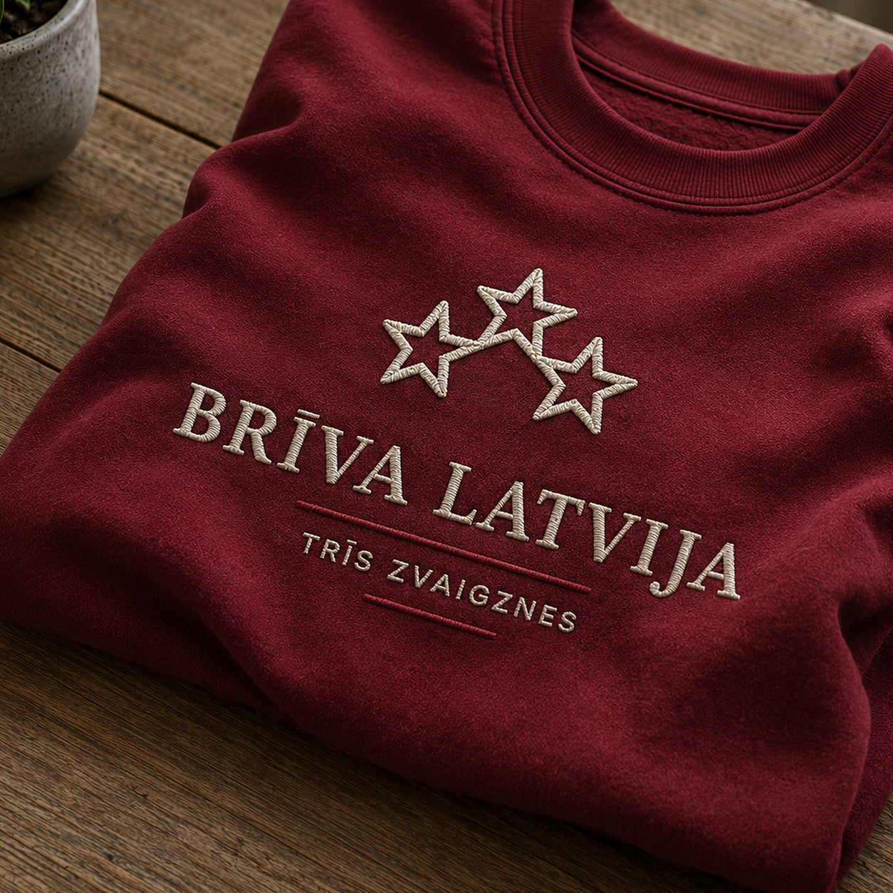 Tuvāks “Brīva Latvija” kompozīcijas skats uz bordo salocītu džemperi ar tekstu “Brīva Latvija” un apakštekstu “Trīs zvaigznes”