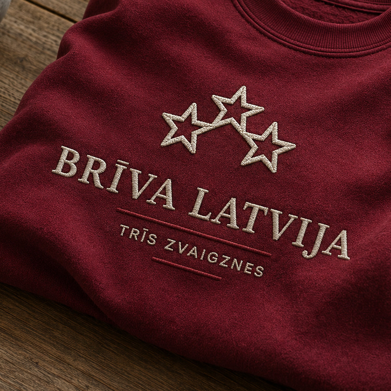 “Brīva Latvija” izšuvuma detaļu tuvplāns uz bordo džempera auduma, redzams motīvs: vārdi Brīva Latvija un trīs zvaigžņu tēls kā valsts novadu vienotības akcents