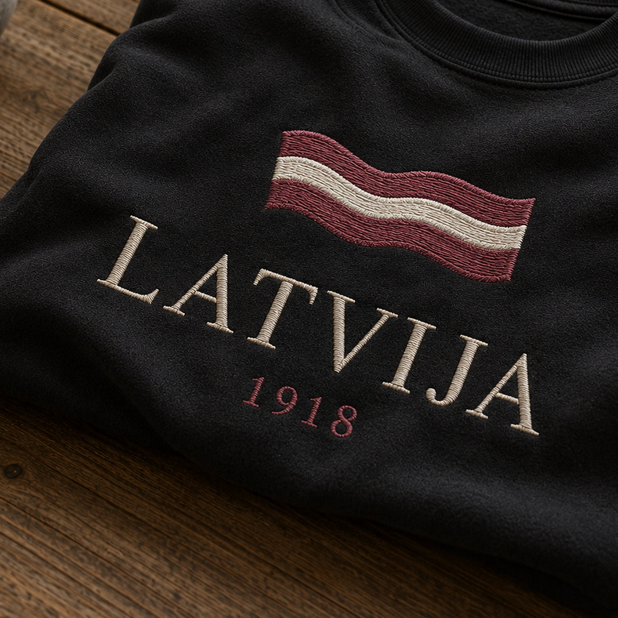 “Latvija 1918” izšuvuma detaļu tuvplāns uz melna džempera auduma, redzams motīvs: Latvijas vārds un vēsturiskais gadskaitlis 1918 tīrā tipogrāfiskā salikumā