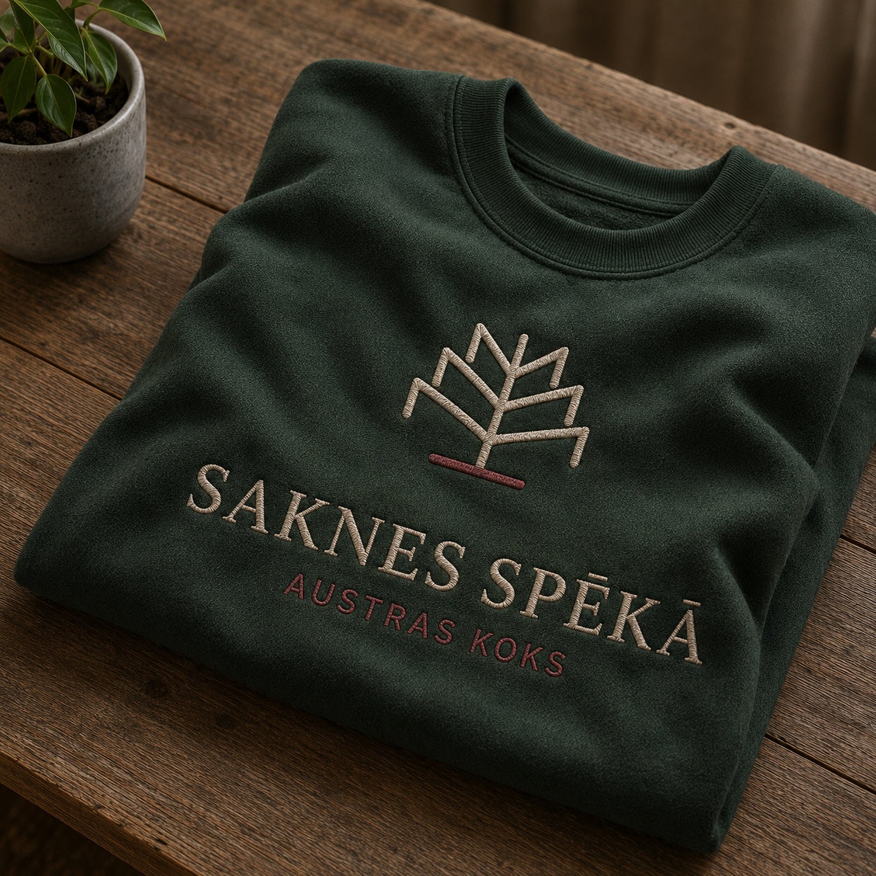 Meža zaļš salocīts džemperis ar “Saknes spēkā” izšuvumu plašākā plakanā izkārtojuma kopskatā