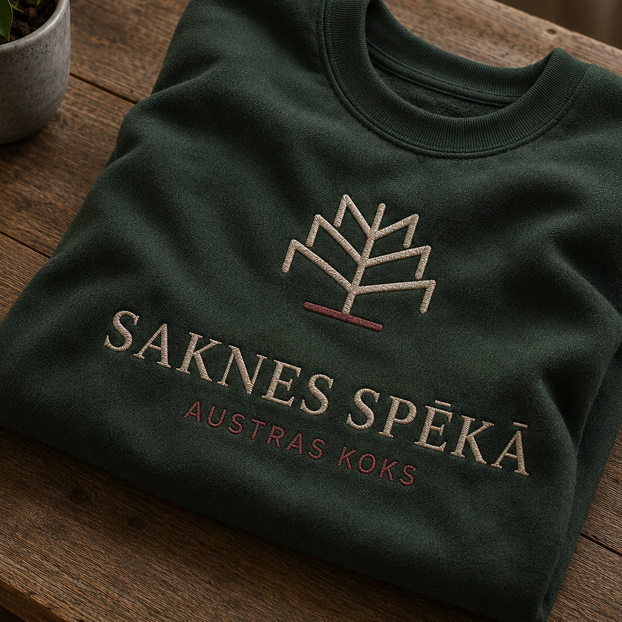 Tuvāks “Saknes spēkā” kompozīcijas skats uz meža zaļu salocītu džemperi ar tekstu “Saknes spēkā” un apakštekstu “Austras koks”