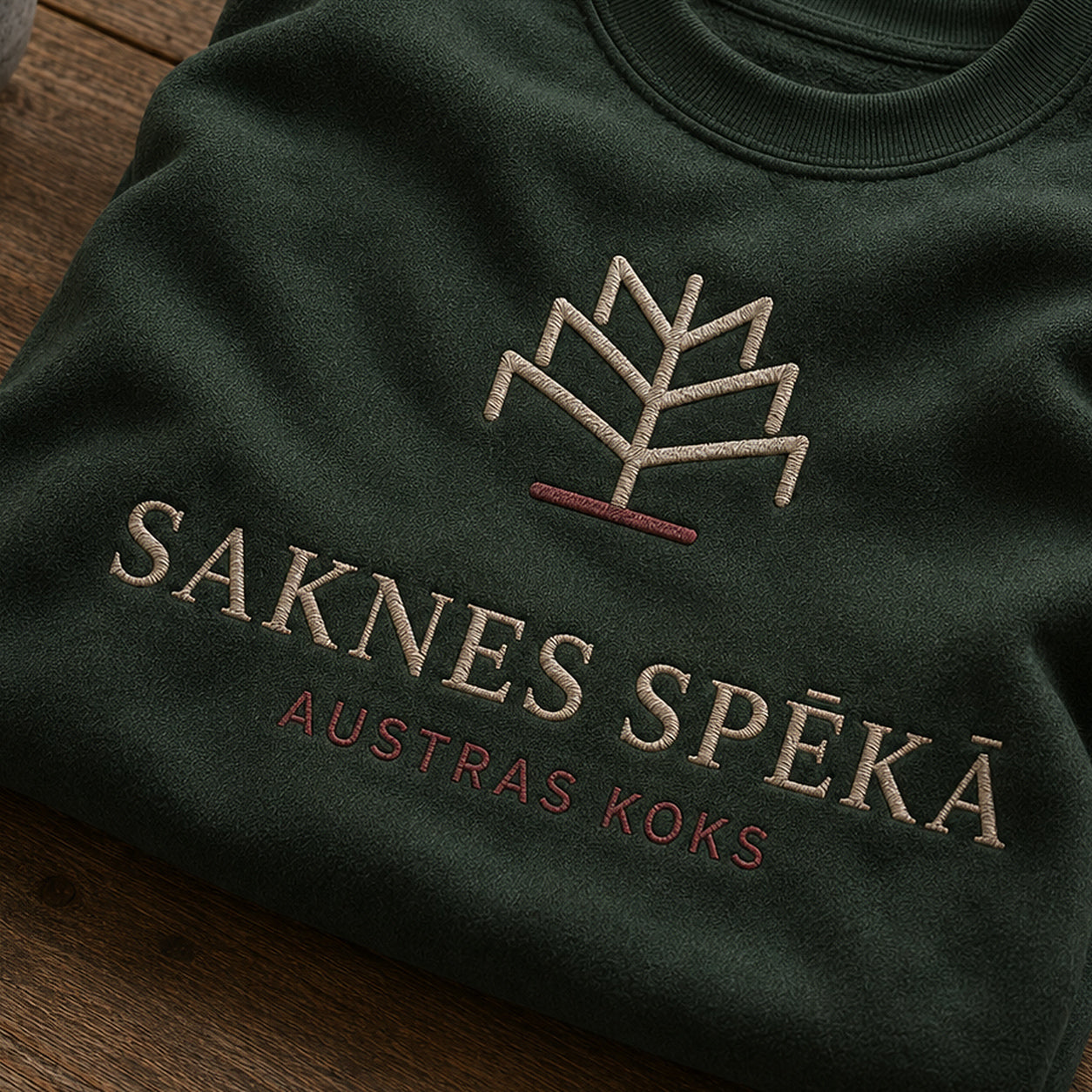“Saknes spēkā” izšuvuma detaļu tuvplāns uz meža zaļa džempera auduma, redzams motīvs: Austras koks ar vēstījumu Saknes spēkā