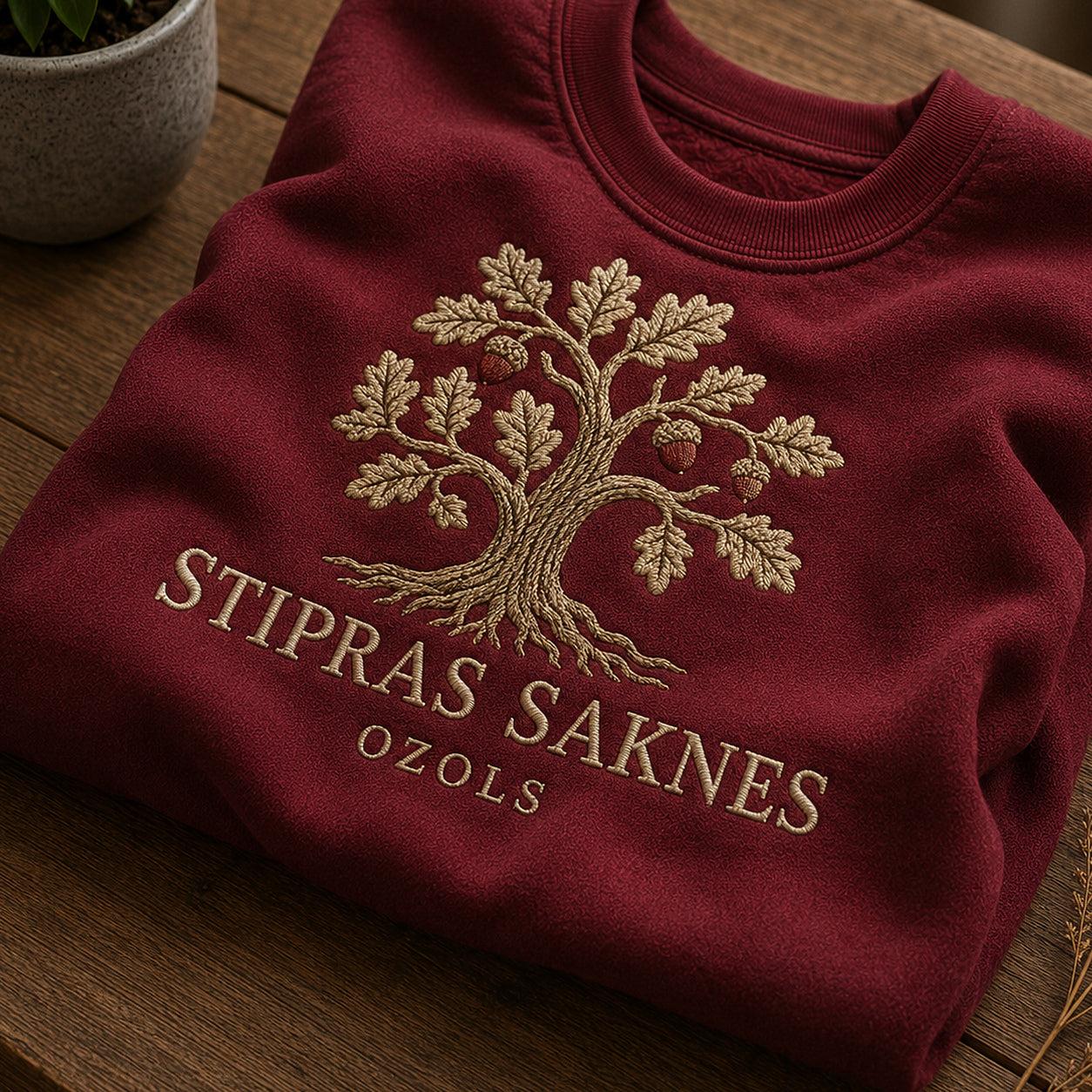 Tuvāks “Stipras saknes” kompozīcijas skats uz bordo salocītu džemperi ar tekstu “Stipras saknes” un apakštekstu “Ozols”