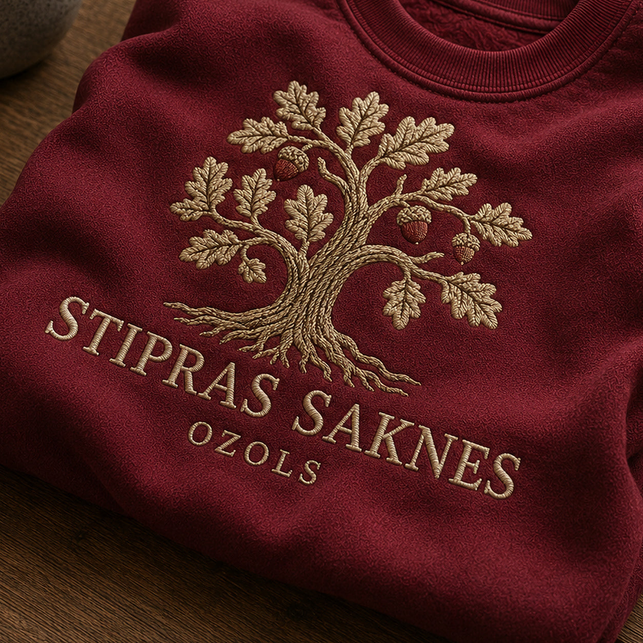 “Stipras saknes” izšuvuma detaļu tuvplāns uz bordo džempera auduma, redzams motīvs: ozola siluets un uzraksts Stipras saknes