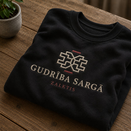 Melns salocīts džemperis ar “Gudrība sargā” izšuvumu plašākā plakanā izkārtojuma kopskatā