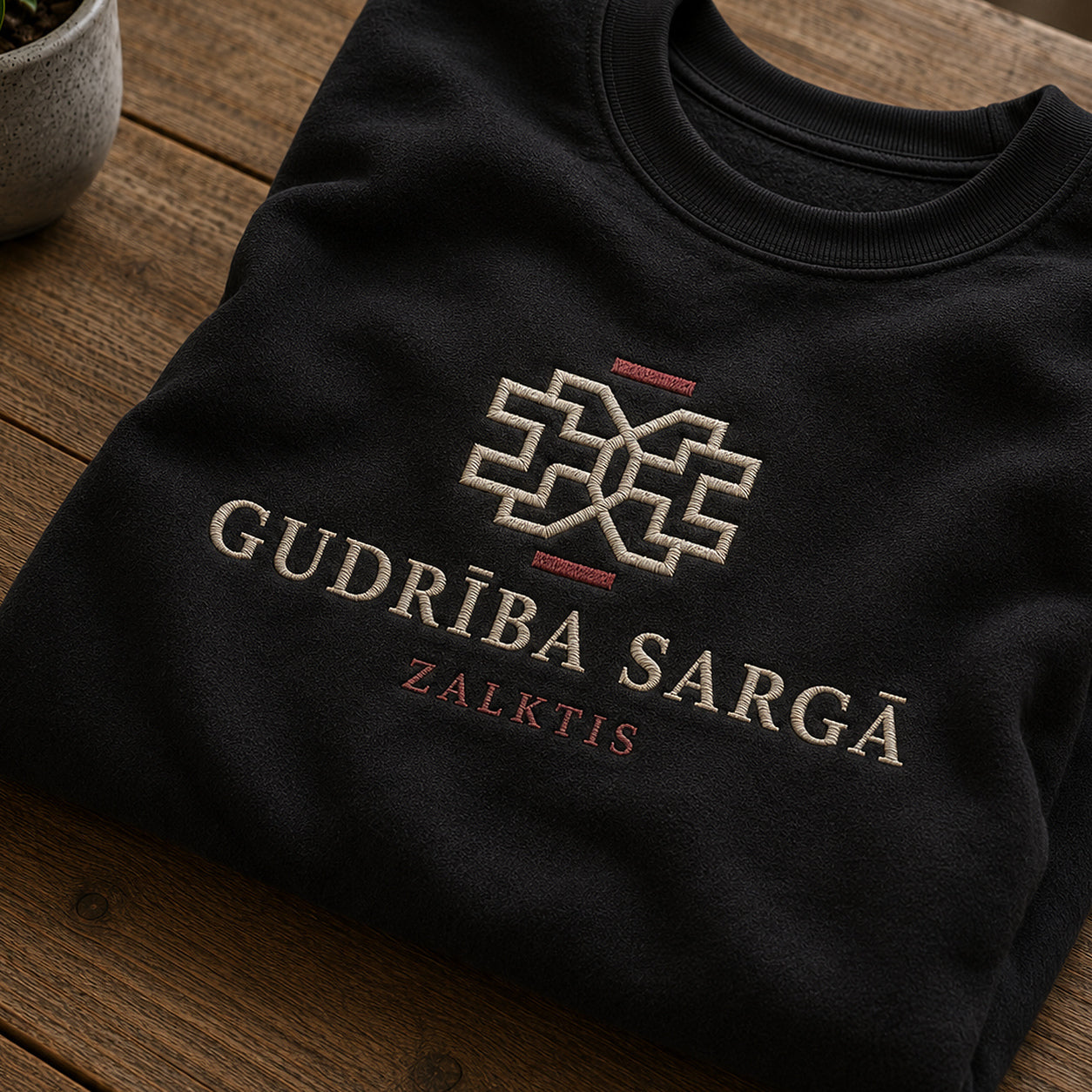 Tuvāks “Gudrība sargā” kompozīcijas skats uz melnu salocītu džemperi ar tekstu “Gudrība sargā” un apakštekstu “Zalktis”