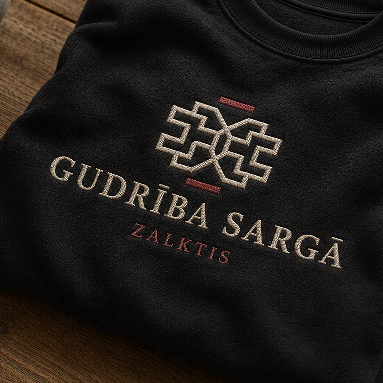 “Gudrība sargā” izšuvuma detaļu tuvplāns uz melna džempera auduma, redzams motīvs: Zalkša līnija un vēstījums Gudrība sargā