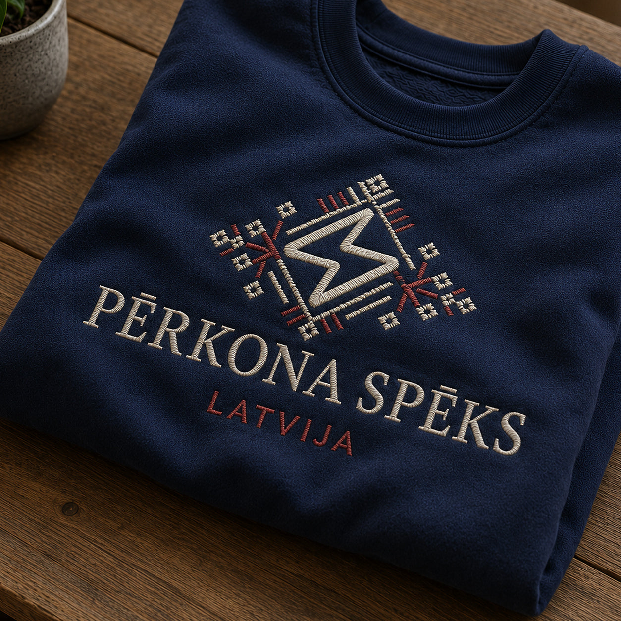 Tuvāks “Pērkona spēks” kompozīcijas skats uz tumši zilu salocītu džemperi ar tekstu “Pērkona spēks” un apakštekstu “Latvija”