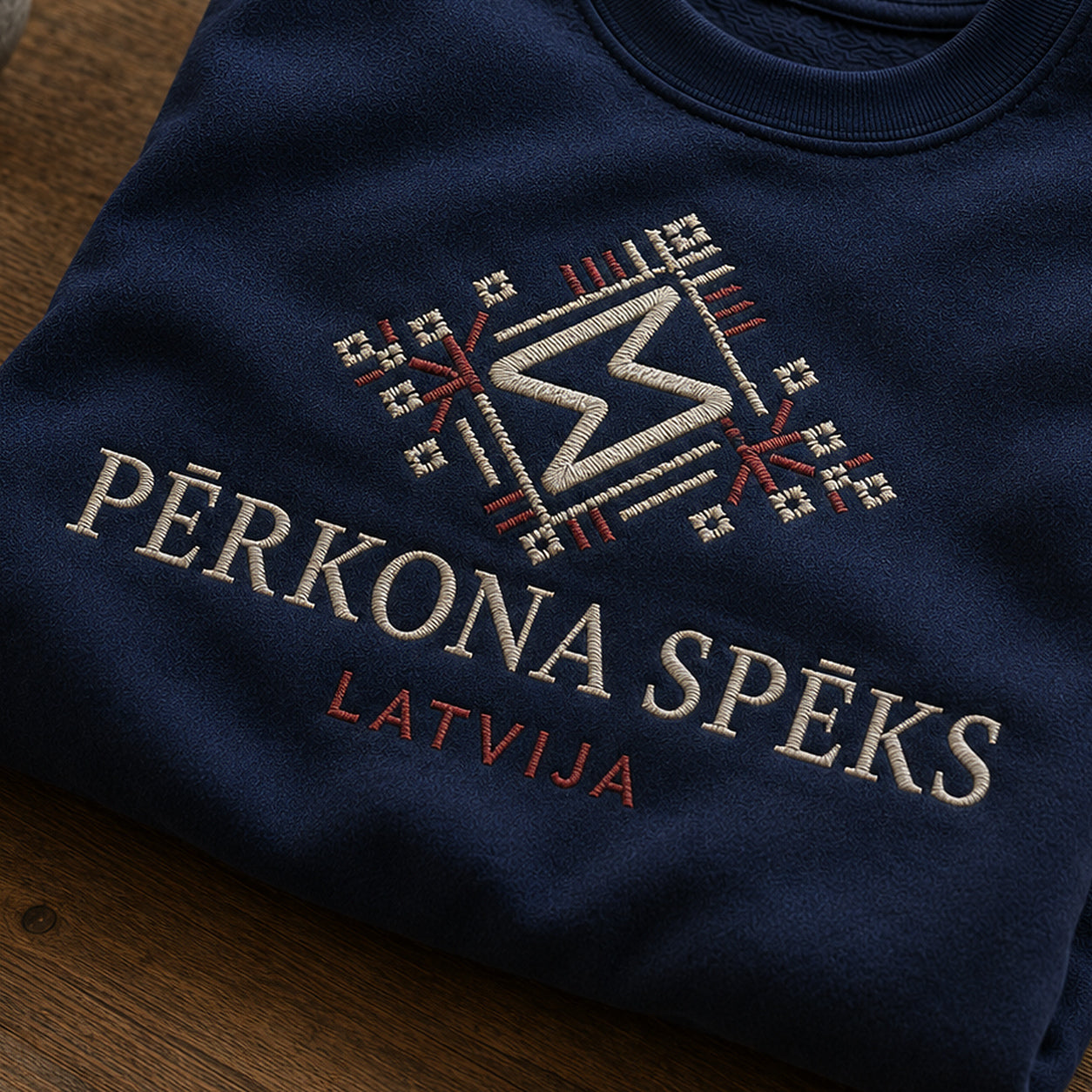 “Pērkona spēks” izšuvuma detaļu tuvplāns uz tumši zila džempera auduma, redzams motīvs: dinamisks Pērkona zīmes raksts kopā ar vārdiem Pērkona spēks