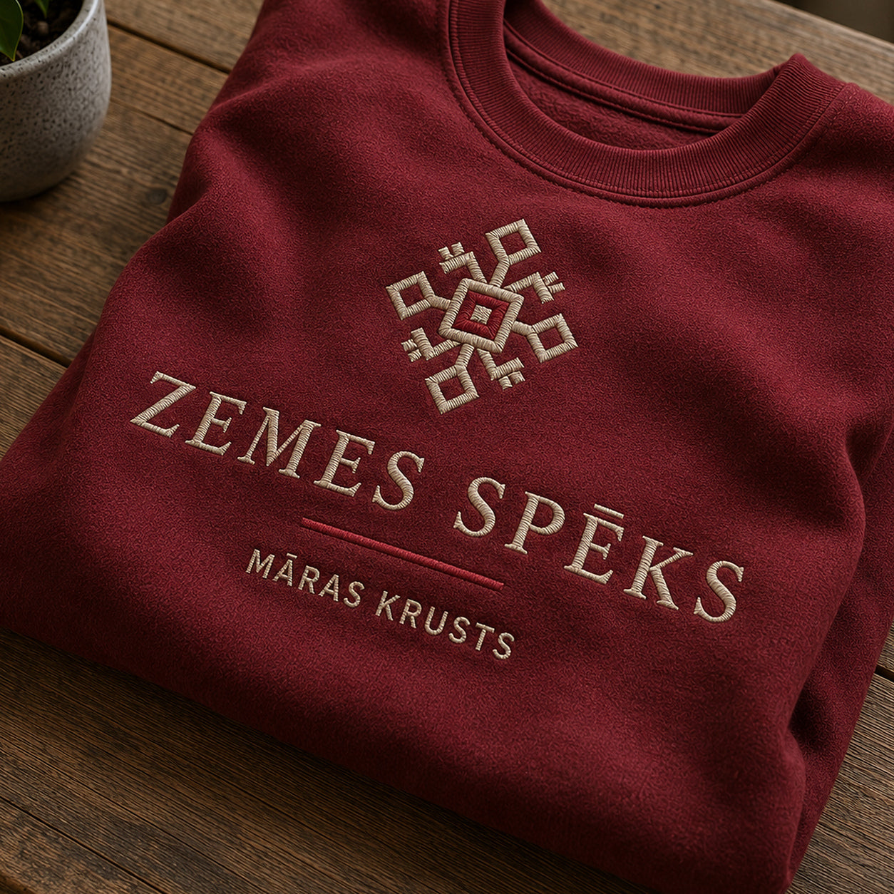 Tuvāks “Zemes spēks” kompozīcijas skats uz bordo salocītu džemperi ar tekstu “Zemes spēks” un apakštekstu “Māras krusts”