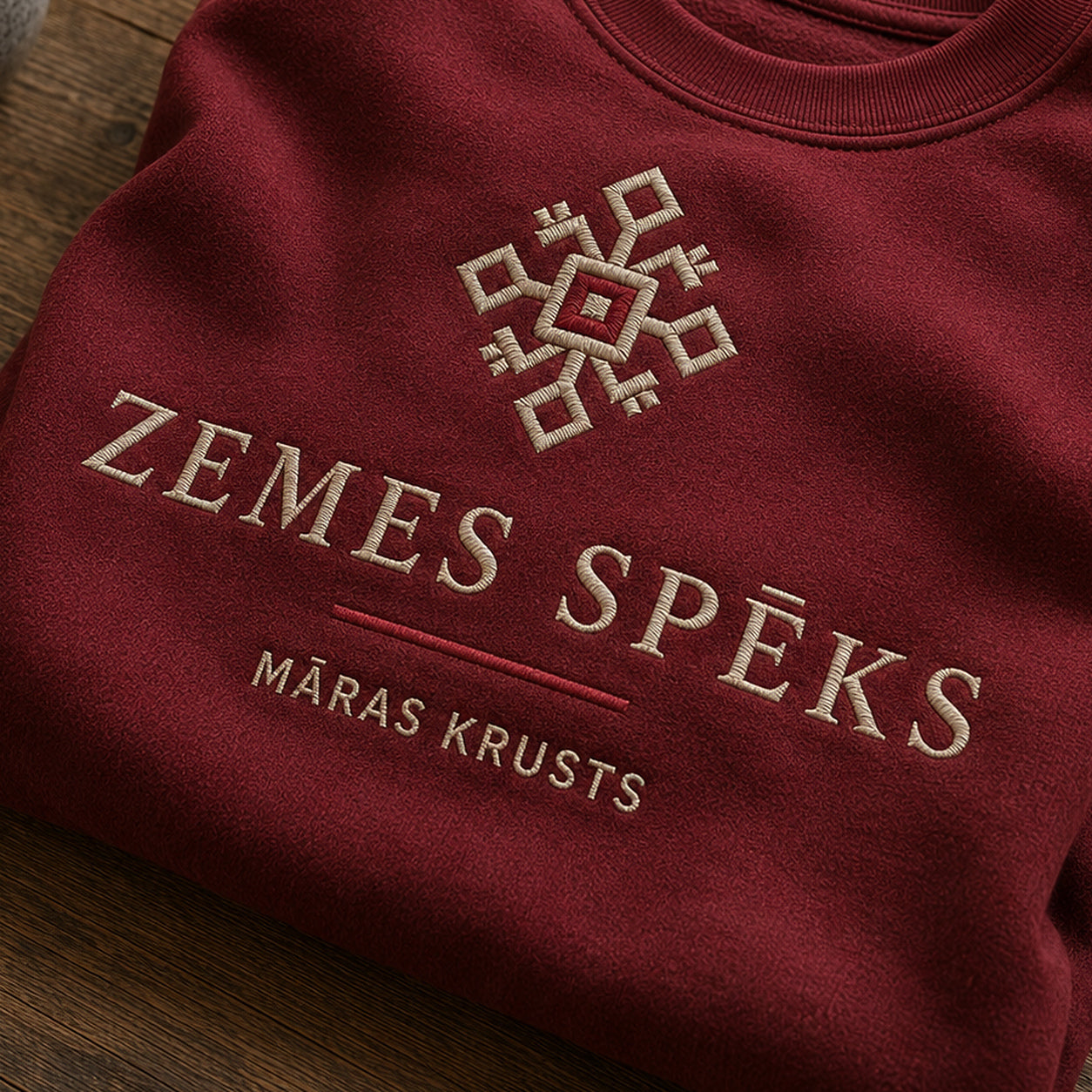 “Zemes spēks” izšuvuma detaļu tuvplāns uz bordo džempera auduma, redzams motīvs: Māras krusta ģeometrija un zemes stabilitātes vēstījums