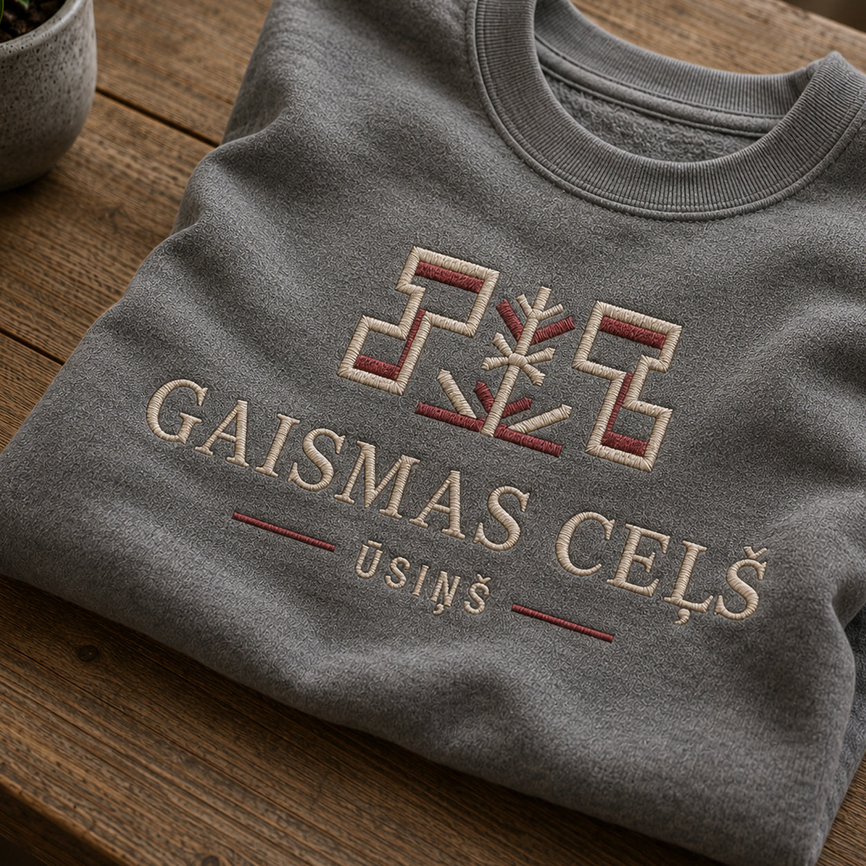 Tuvāks “Gaismas ceļš” kompozīcijas skats uz pelēkas melanžas salocītu džemperi ar tekstu “Gaismas ceļš” un apakštekstu “Ūsiņš”
