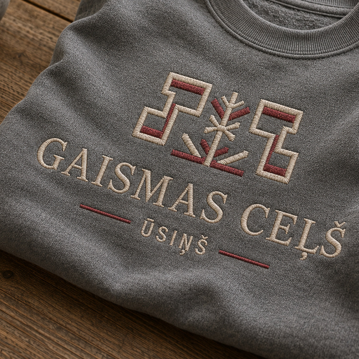 “Gaismas ceļš” izšuvuma detaļu tuvplāns uz pelēkas melanžas džempera auduma, redzams motīvs: Ūsiņa zīme un virzošais uzraksts Gaismas ceļš