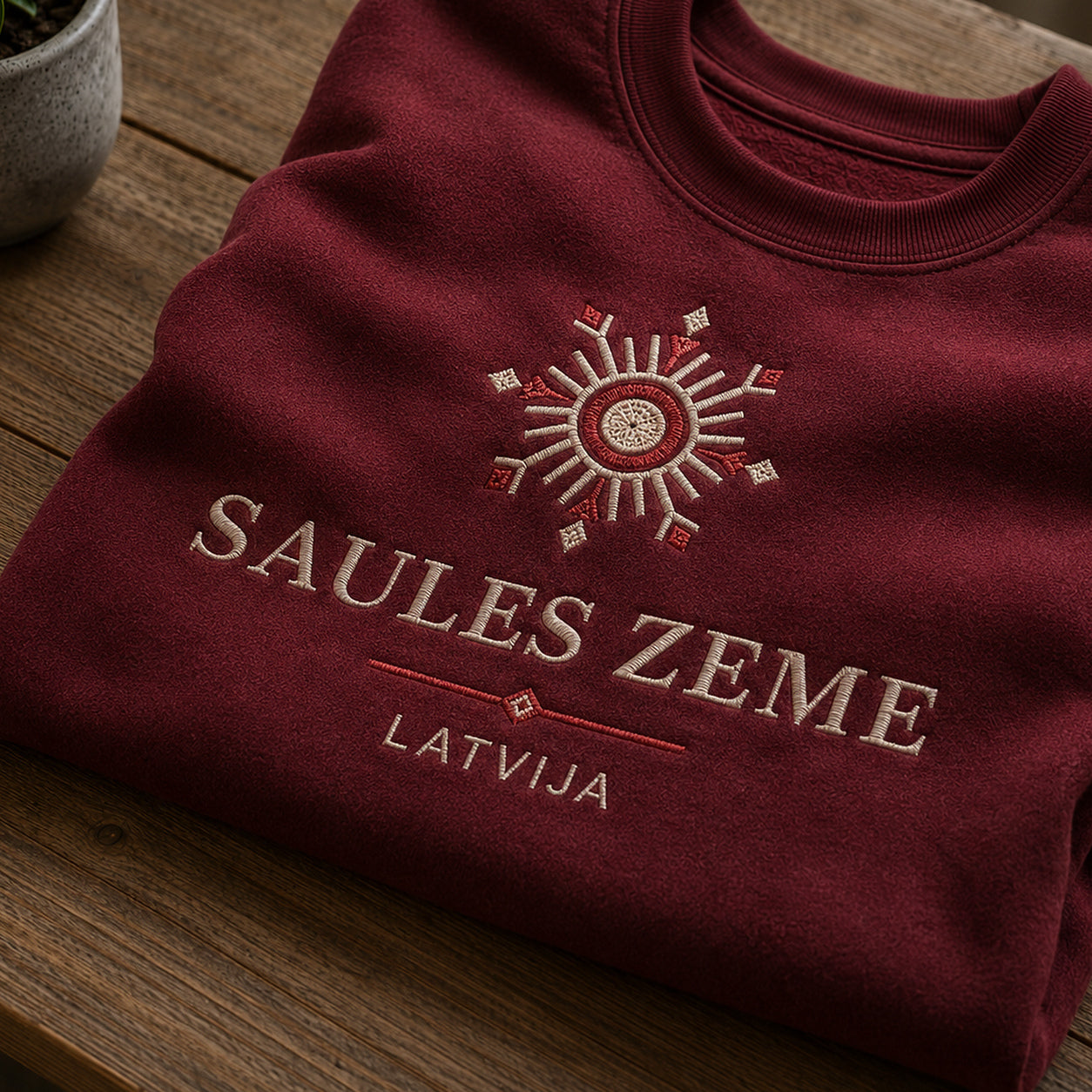 Tuvāks “Saules zeme Latvija” kompozīcijas skats uz bordo salocītu džemperi ar tekstu “Saules zeme” un apakštekstu “Latvija”