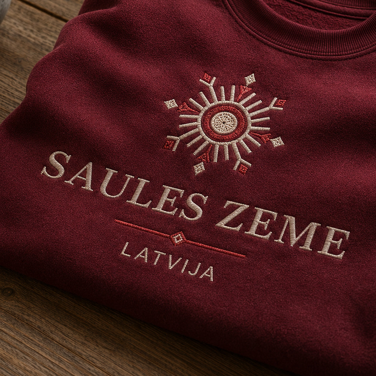 “Saules zeme Latvija” izšuvuma detaļu tuvplāns uz bordo džempera auduma, redzams motīvs: saules ritms kopā ar vārdiem Saules zeme Latvija