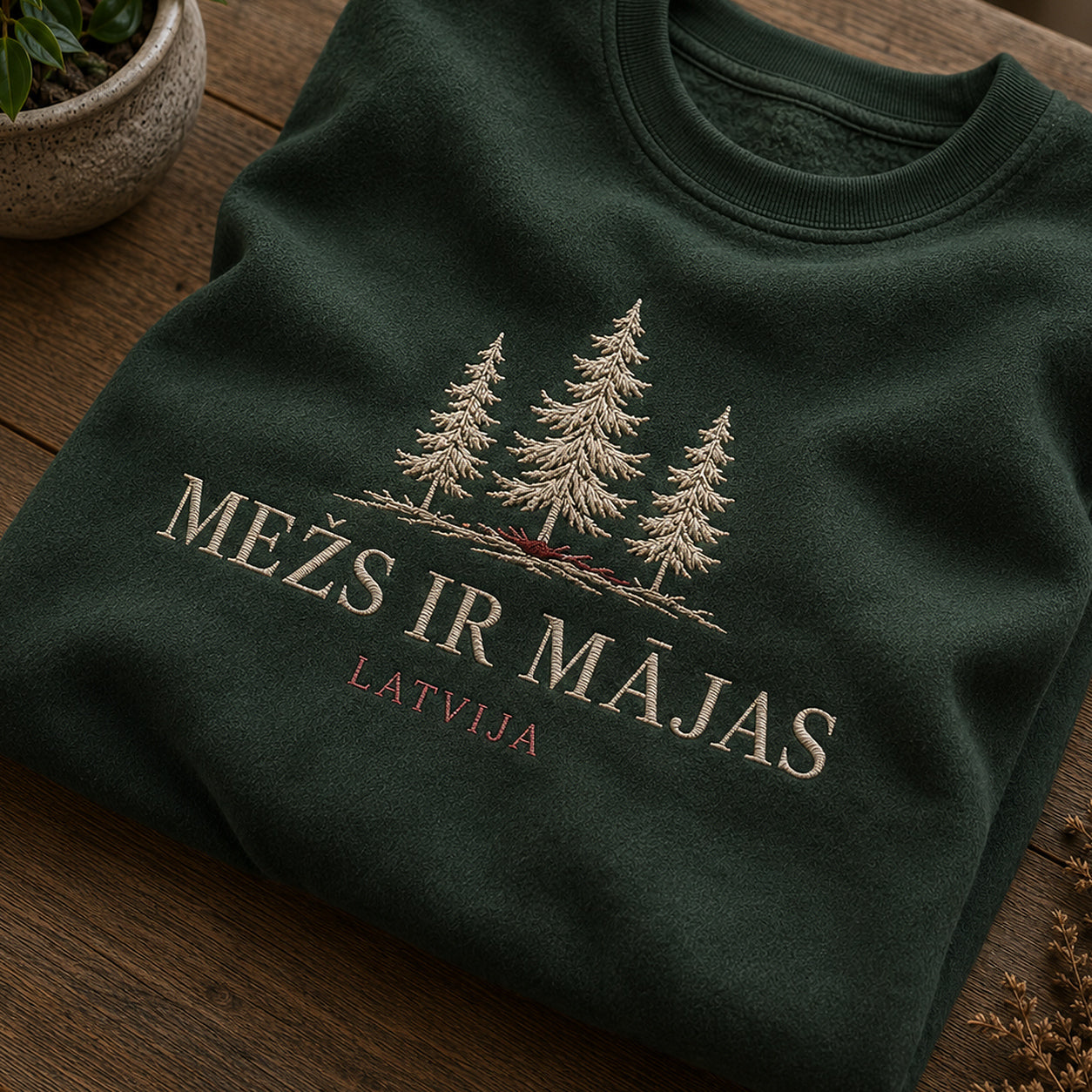 Tuvāks “Mežs ir mājas” kompozīcijas skats uz meža zaļu salocītu džemperi ar tekstu “Mežs ir mājas” un apakštekstu “Latvija”