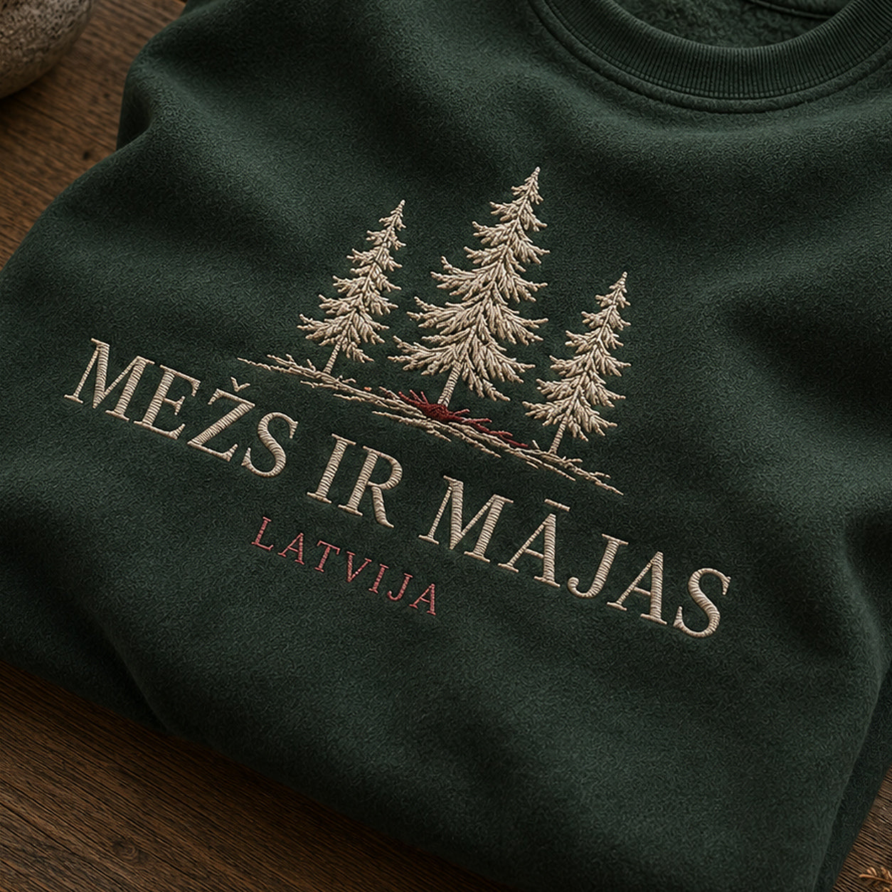 “Mežs ir mājas” izšuvuma detaļu tuvplāns uz meža zaļa džempera auduma, redzams motīvs: vārdi Mežs ir mājas kopā ar tiešu norādi uz Latviju