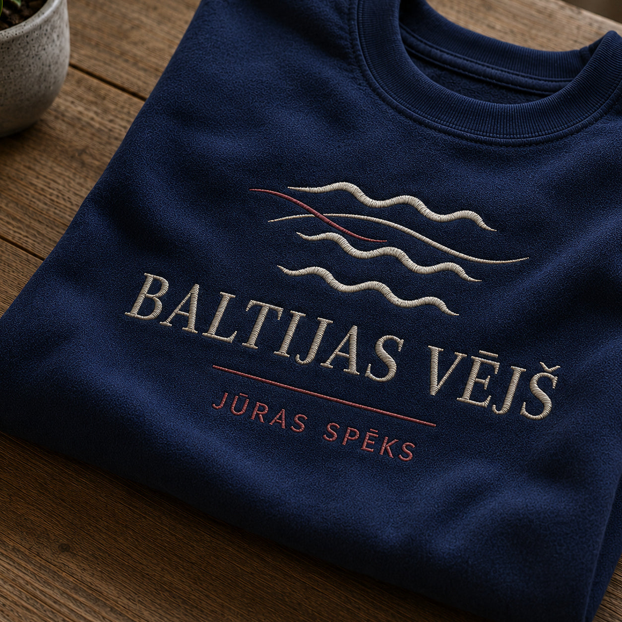Tuvāks “Baltijas vējš” kompozīcijas skats uz tumši zilu salocītu džemperi ar tekstu “Baltijas vējš” un apakštekstu “Jūras spēks”