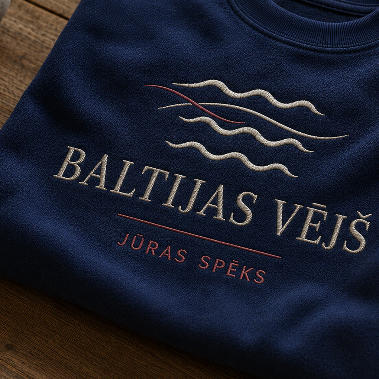 “Baltijas vējš” izšuvuma detaļu tuvplāns uz tumši zila džempera auduma, redzams motīvs: plūstošs Baltijas vēja tēls un vārdi Jūras spēks
