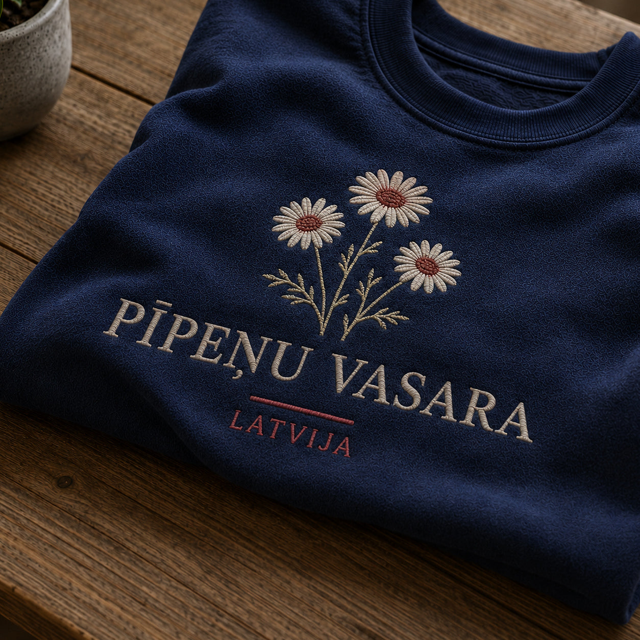 Tuvāks “Pīpeņu vasara” kompozīcijas skats uz tumši zilu salocītu džemperi ar tekstu “Pīpeņu vasara” un apakštekstu “Latvija”