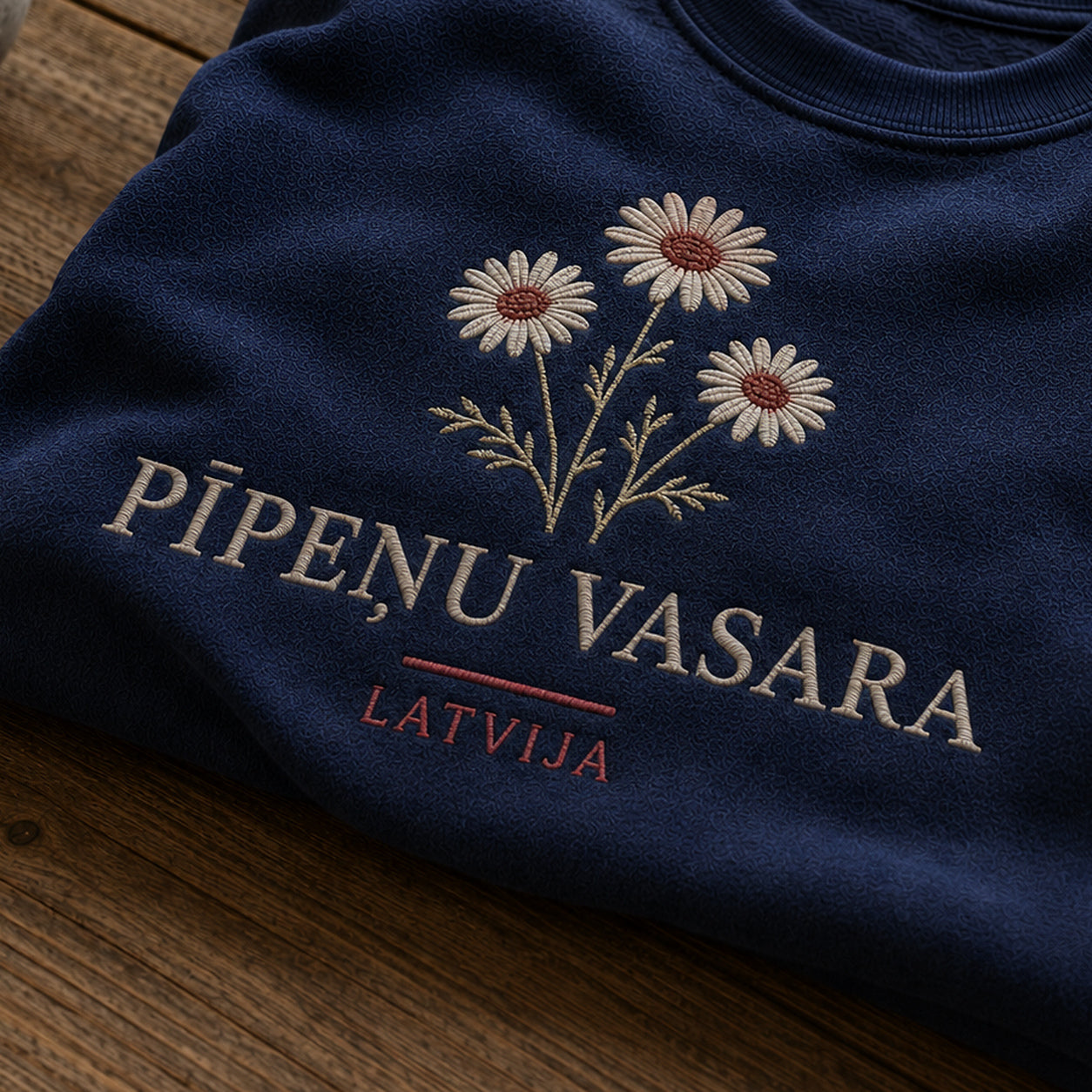 “Pīpeņu vasara” izšuvuma detaļu tuvplāns uz tumši zila džempera auduma, redzams motīvs: pīpeņu ziedu vieglums un vārdi Pīpeņu vasara Latvija