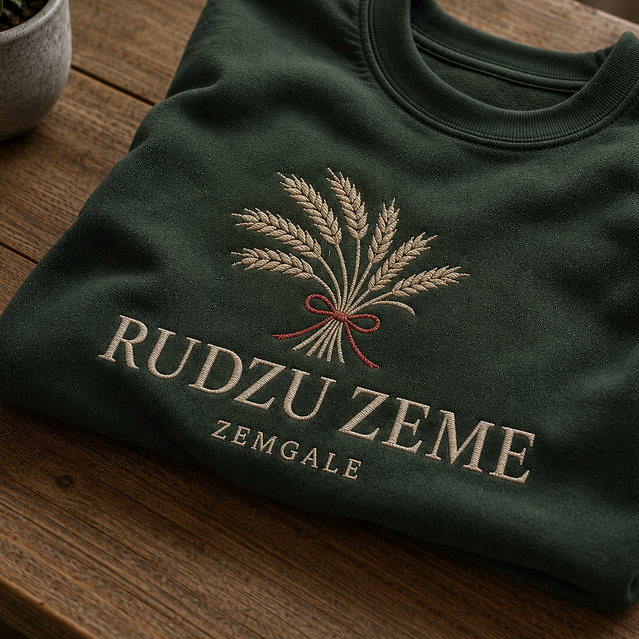 Tuvāks “Rudzu zeme” kompozīcijas skats uz meža zaļu salocītu džemperi ar tekstu “Rudzu zeme” un apakštekstu “Zemgale”
