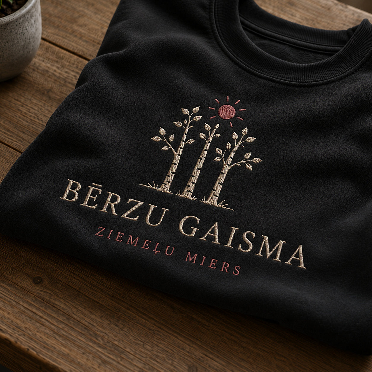 Tuvāks “Bērzu gaisma” kompozīcijas skats uz melnu salocītu džemperi ar tekstu “Bērzu gaisma” un apakštekstu “Ziemeļu miers”