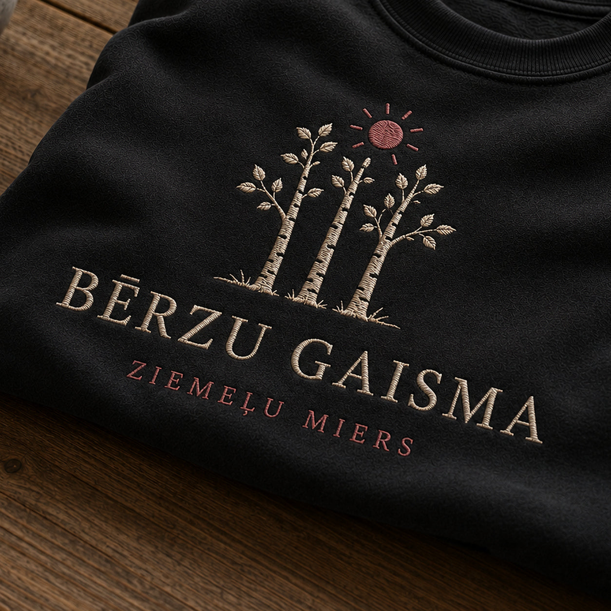 “Bērzu gaisma” izšuvuma detaļu tuvplāns uz melna džempera auduma, redzams motīvs: bērzu gaismas kontrasts un mierīgais uzraksts Ziemeļu miers