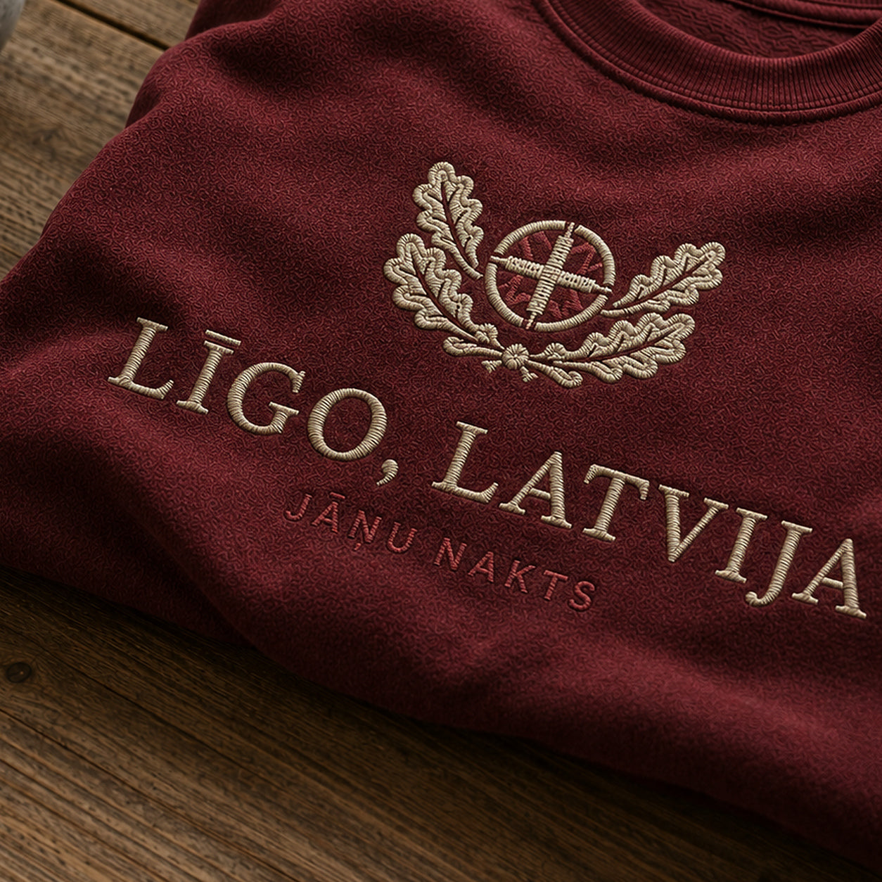 “Līgo, Latvija” izšuvuma detaļu tuvplāns uz bordo džempera auduma, redzams motīvs: svinīgais aicinājums Līgo, Latvija un Jāņu nakts noskaņa