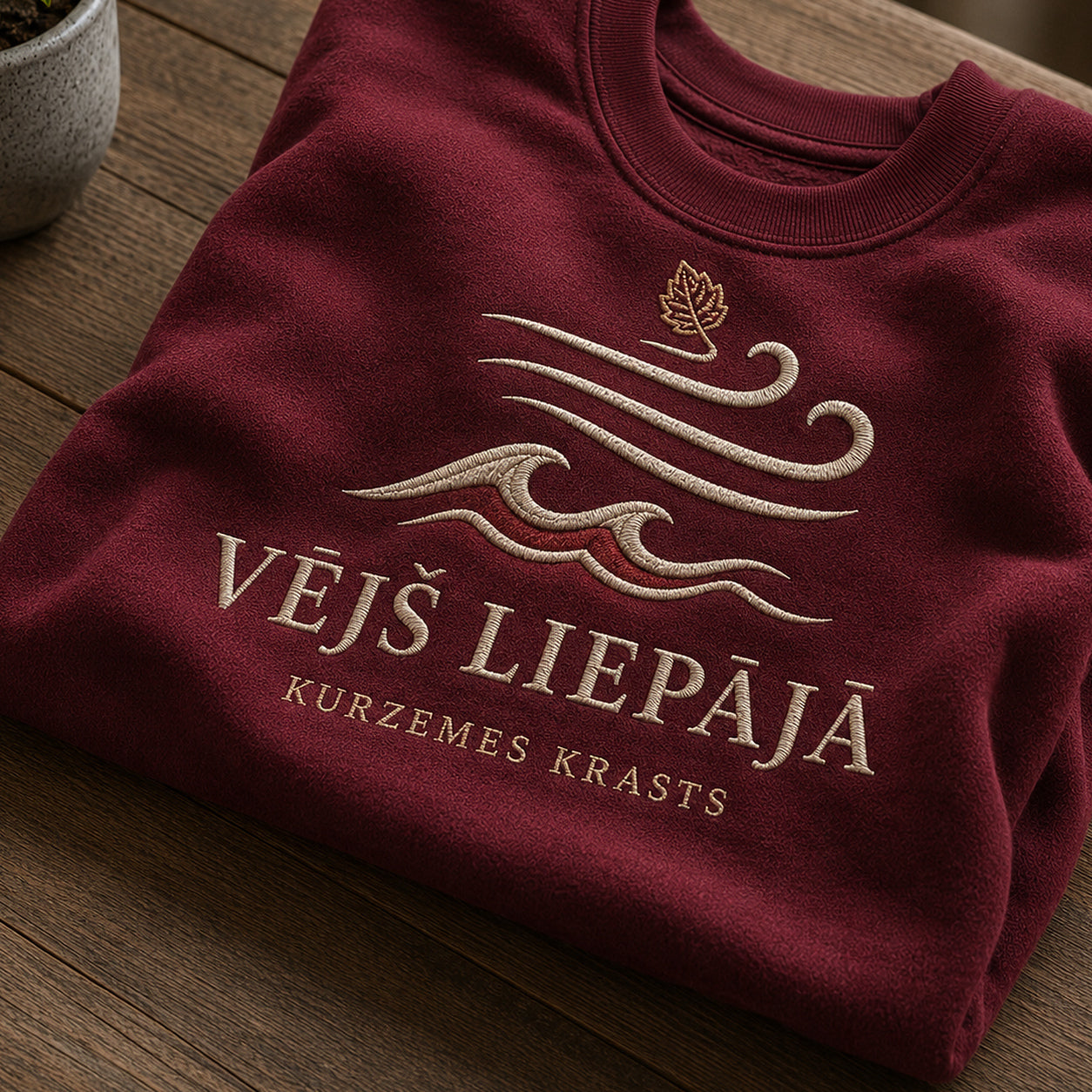 Tuvāks “Vējš Liepājā” kompozīcijas skats uz bordo salocītu džemperi ar tekstu “Vējš Liepājā” un apakštekstu “Kurzemes krasts”