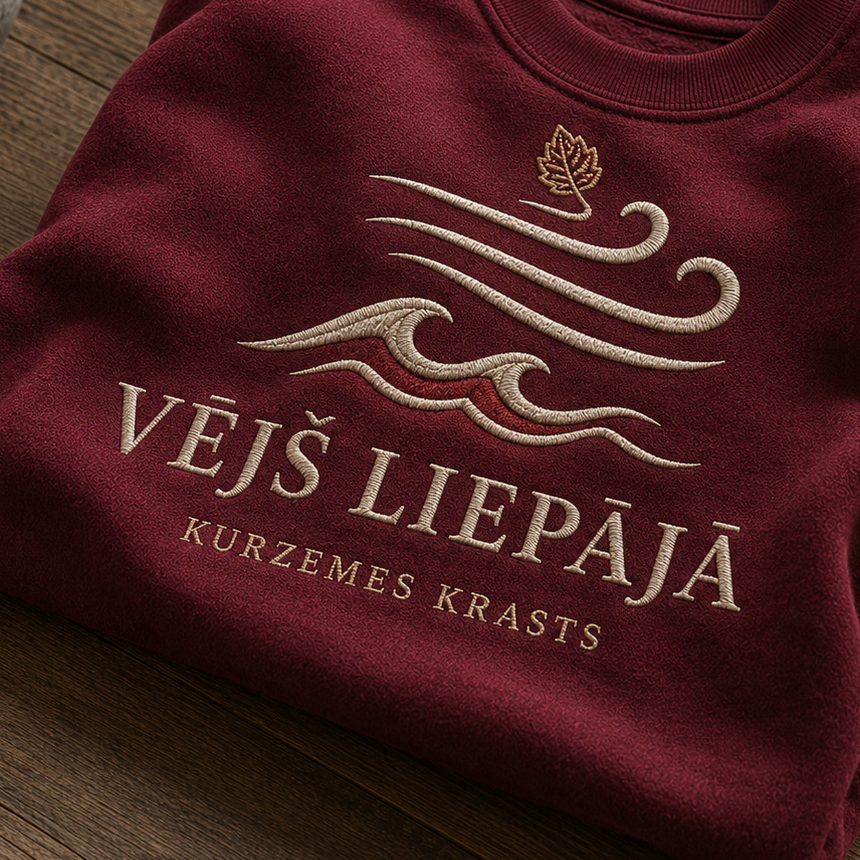 “Vējš Liepājā” izšuvuma detaļu tuvplāns uz bordo džempera auduma, redzams motīvs: kustīgs uzraksts Vējš Liepājā un Kurzemes krasta sajūta