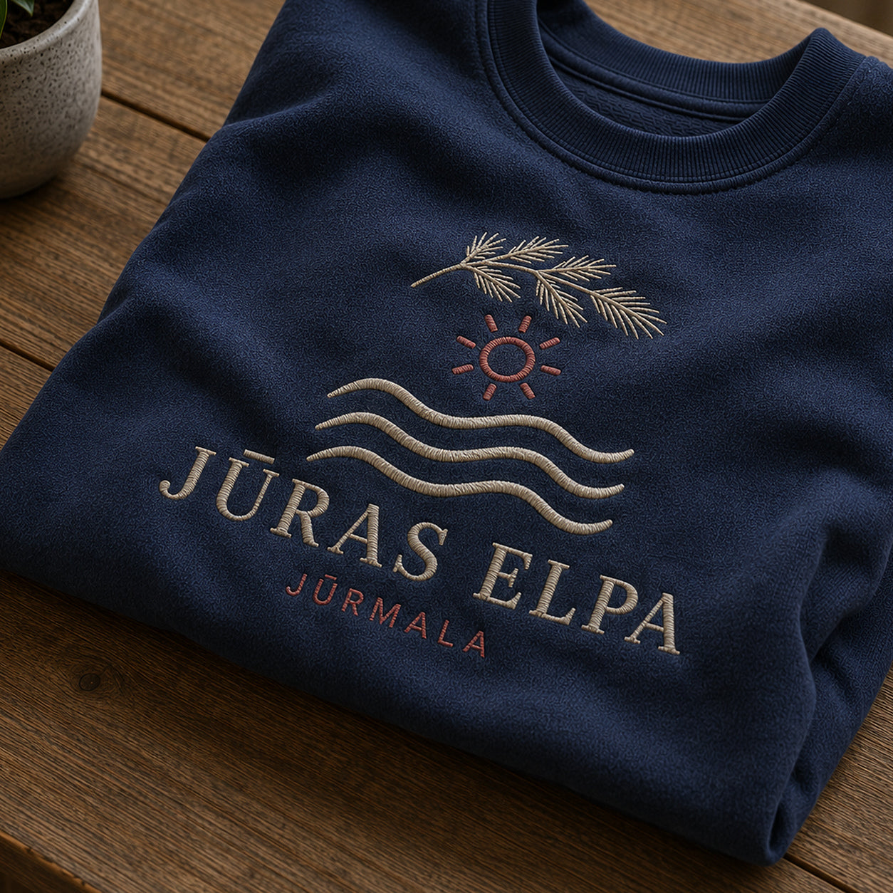 Tuvāks “Jūras elpa” kompozīcijas skats uz tumši zilu salocītu džemperi ar tekstu “Jūras elpa” un apakštekstu “Jūrmala”