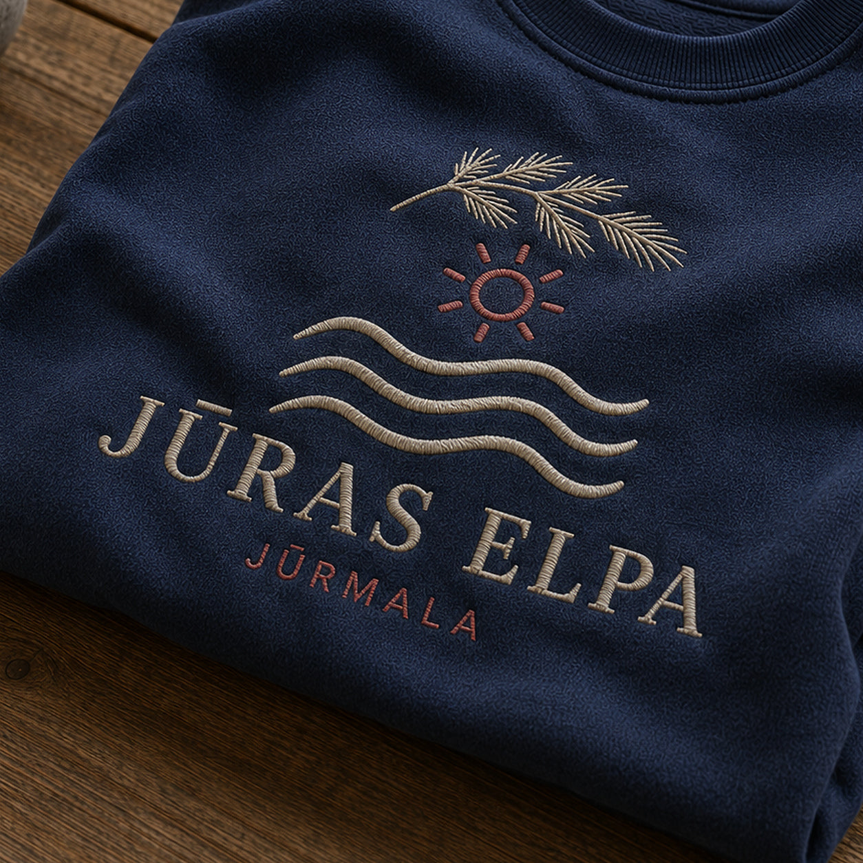 “Jūras elpa” izšuvuma detaļu tuvplāns uz tumši zila džempera auduma, redzams motīvs: Jūras elpas nosaukums un skaidra Jūrmalas piederība