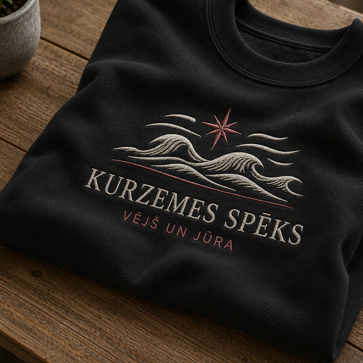 Tuvāks “Kurzemes spēks” kompozīcijas skats uz melnu salocītu džemperi ar tekstu “Kurzemes spēks” un apakštekstu “Vējš un jūra”