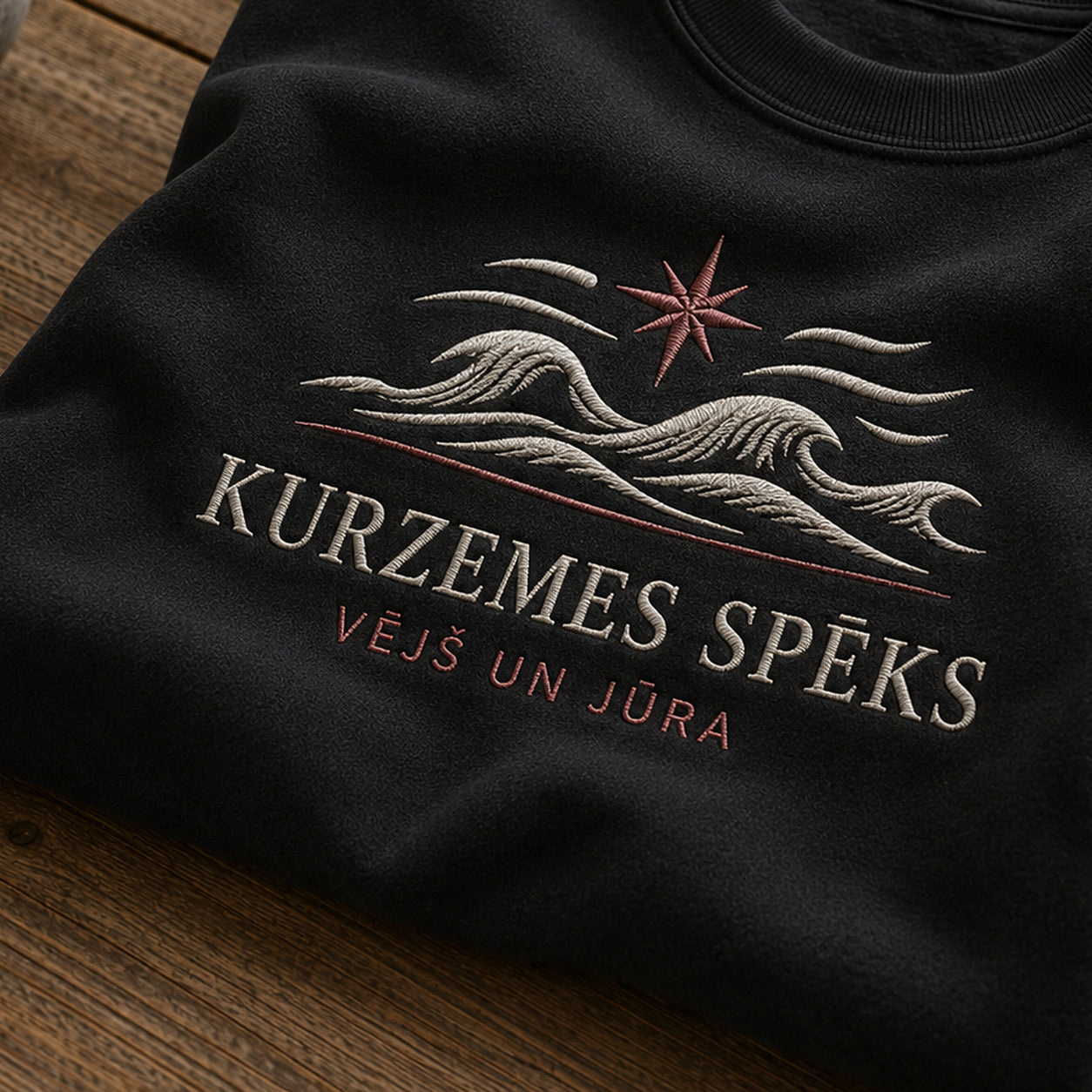 “Kurzemes spēks” izšuvuma detaļu tuvplāns uz melna džempera auduma, redzams motīvs: spēcīgs Kurzemes uzraksts ar vēja un jūras dubulto tēmu