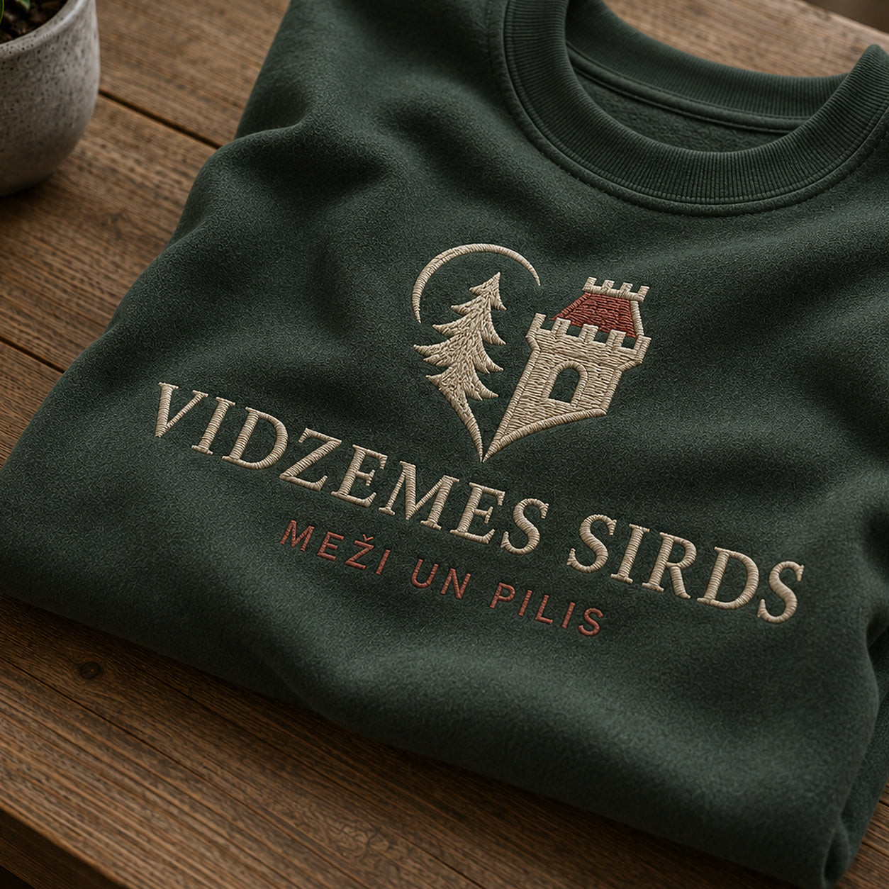 Tuvāks “Vidzemes sirds” kompozīcijas skats uz meža zaļu salocītu džemperi ar tekstu “Vidzemes sirds” un apakštekstu “Meži un pilis”
