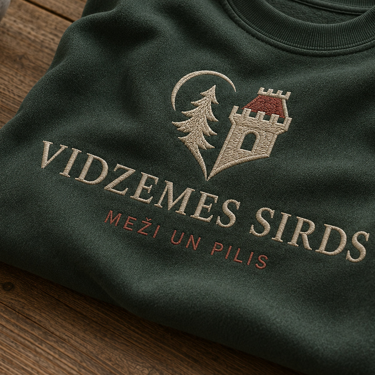 “Vidzemes sirds” izšuvuma detaļu tuvplāns uz meža zaļa džempera auduma, redzams motīvs: Vidzemes sirds ar mežu un piļu stāsta apvienojumu