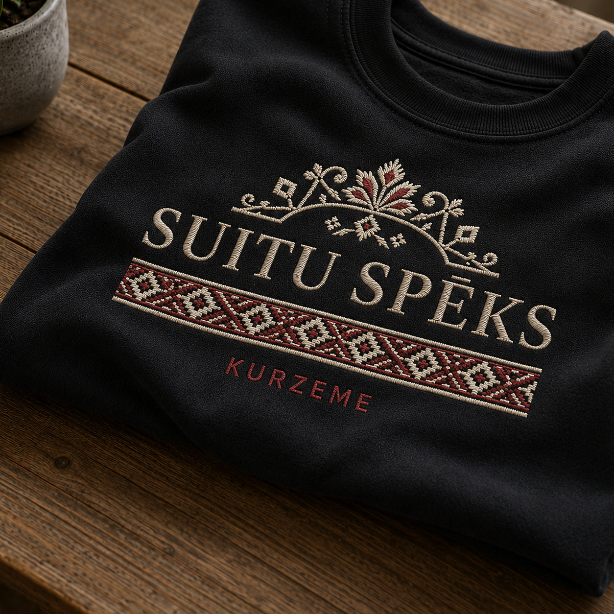 Tuvāks “Suitu spēks” kompozīcijas skats uz melnu salocītu džemperi ar tekstu “Suitu spēks” un apakštekstu “Kurzeme”