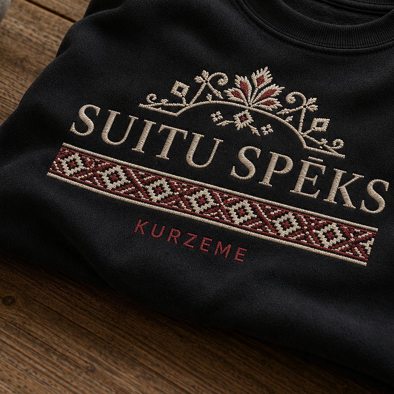 “Suitu spēks” izšuvuma detaļu tuvplāns uz melna džempera auduma, redzams motīvs: Suitu spēka nosaukums un skaidra Kurzemes kultūrtelpas piederība