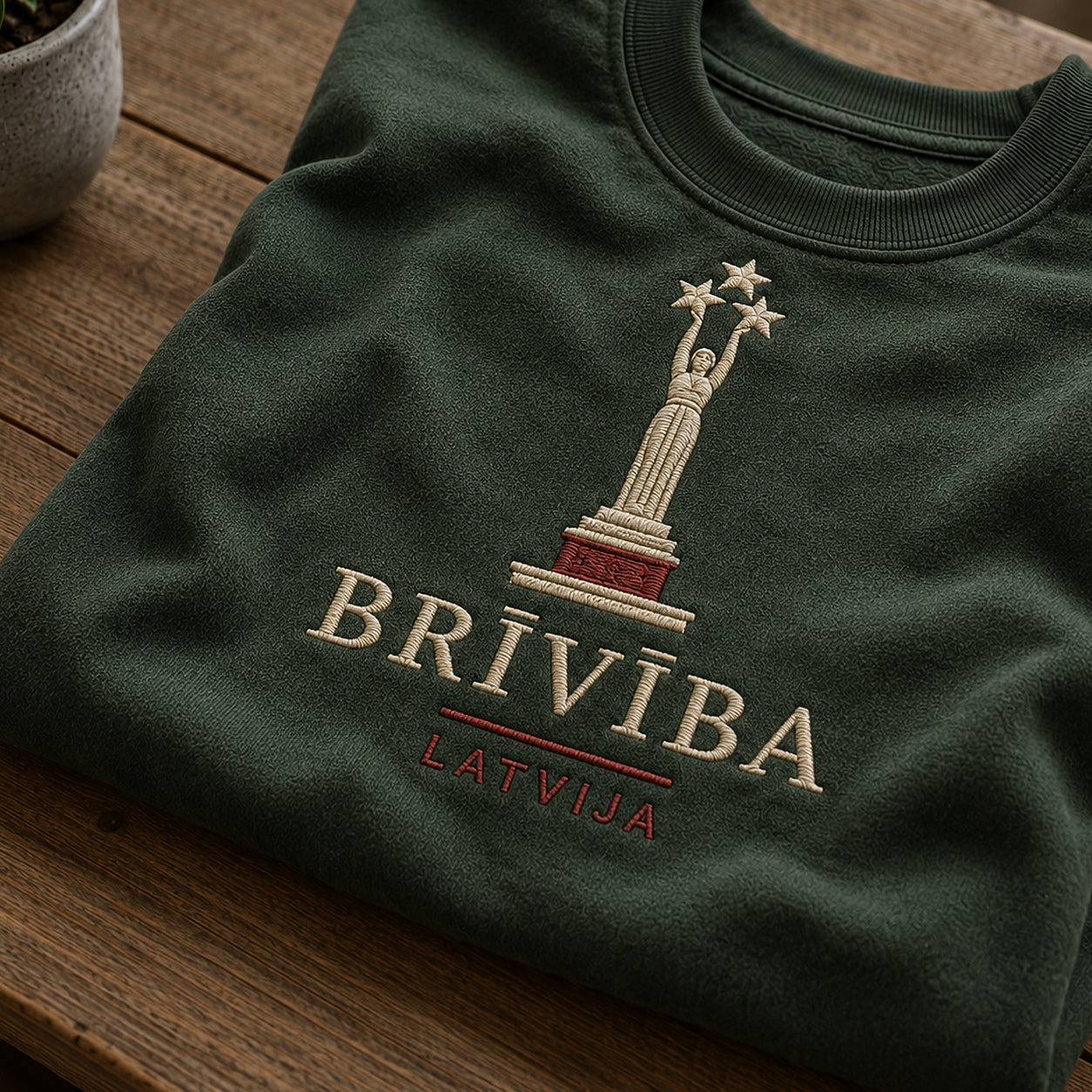 Tuvāks “Brīvība Latvija” kompozīcijas skats uz meža zaļu salocītu džemperi ar tekstu “Brīvība” un apakštekstu “Latvija”