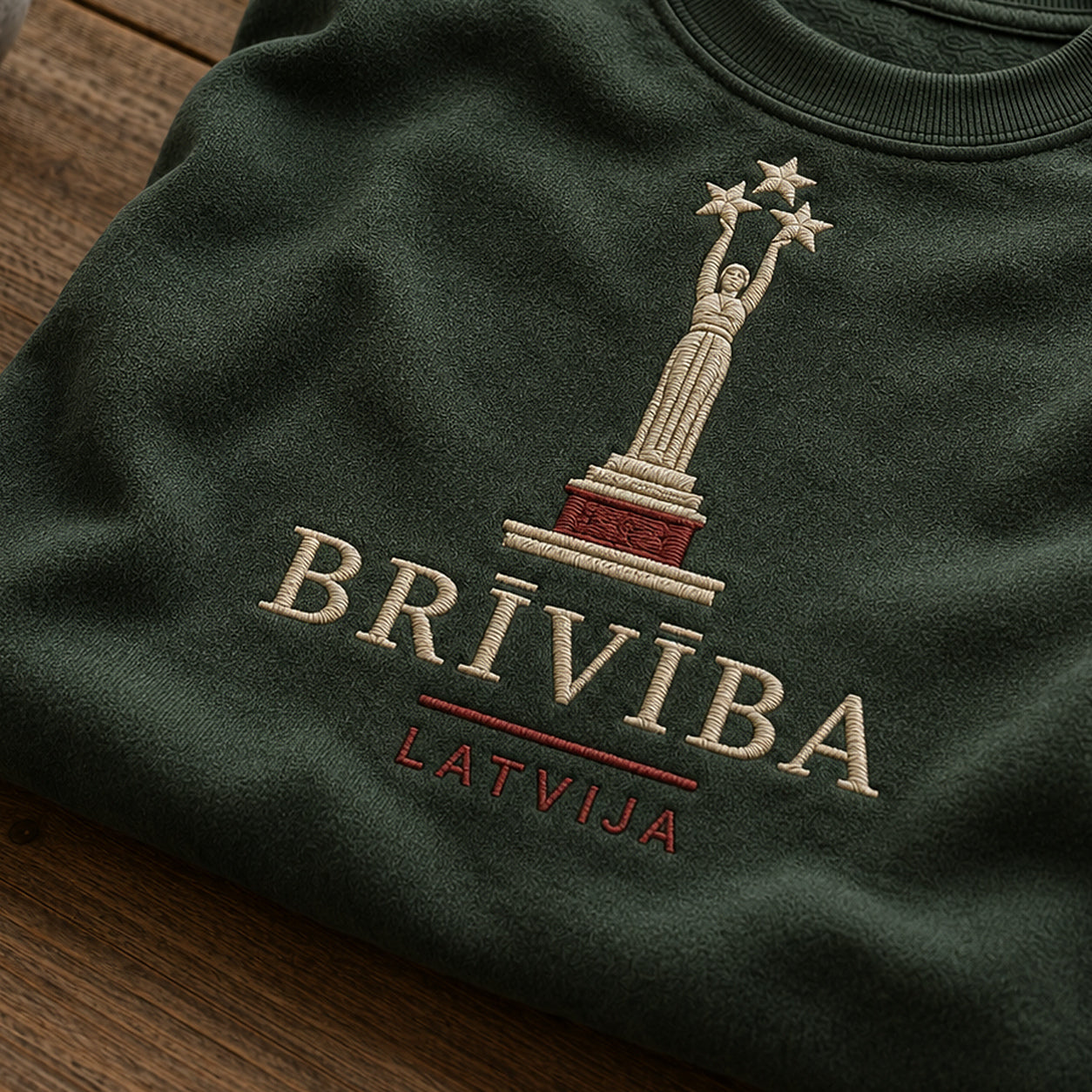 “Brīvība Latvija” izšuvuma detaļu tuvplāns uz meža zaļa džempera auduma, redzams motīvs: viens skaidrs vārds Brīvība un nepārprotama Latvijas piederība