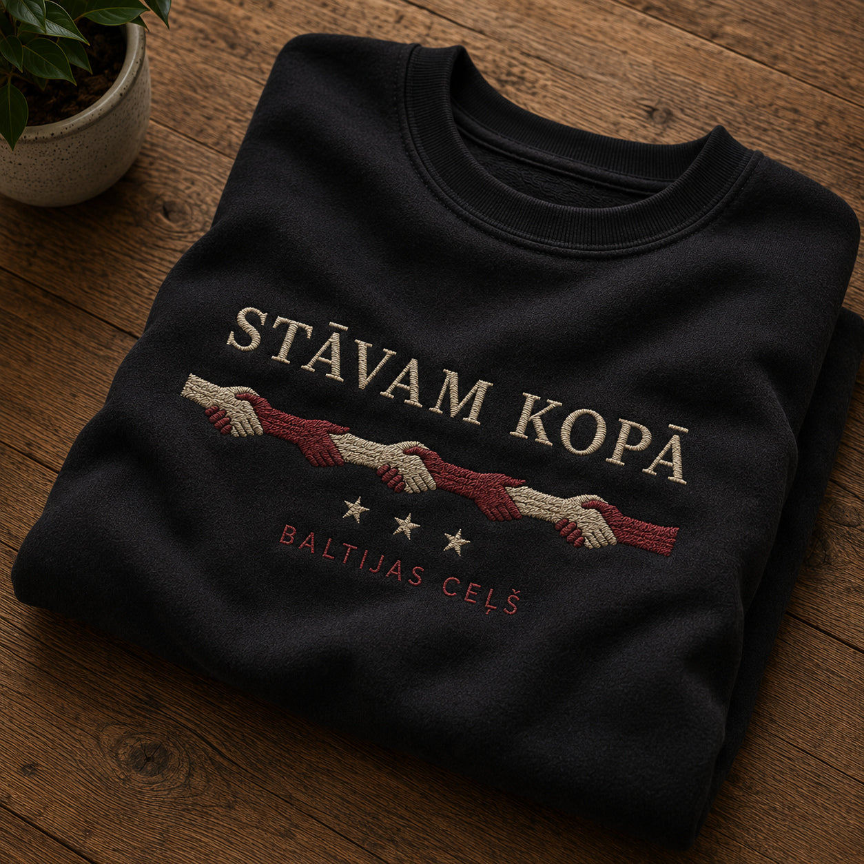 Melns salocīts džemperis ar “Stāvam kopā” izšuvumu plašākā plakanā izkārtojuma kopskatā